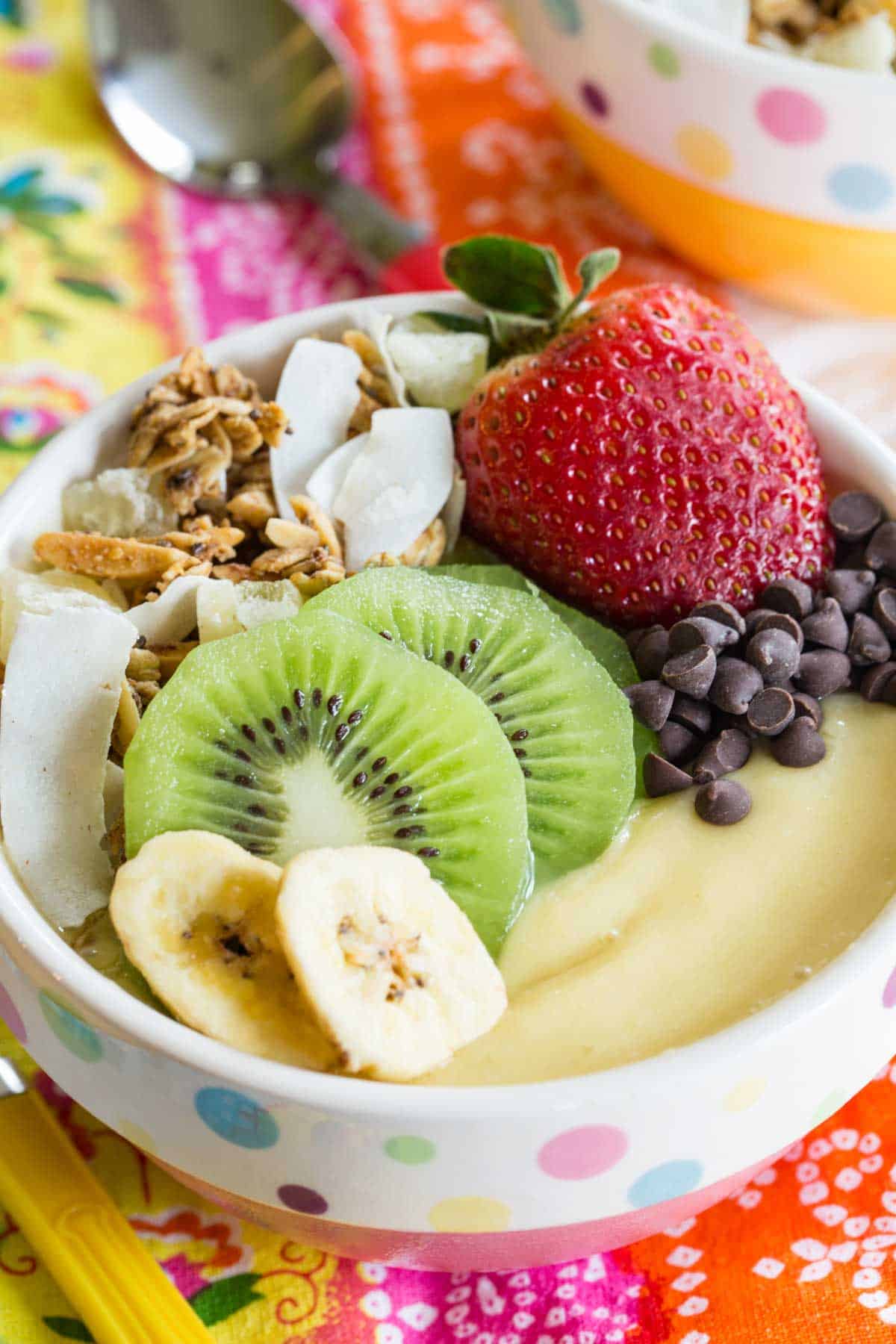 closeup of a pineapple mango smoothie bowl topped with strawberry, kiwi, banana chips, chocolate chips, and granola