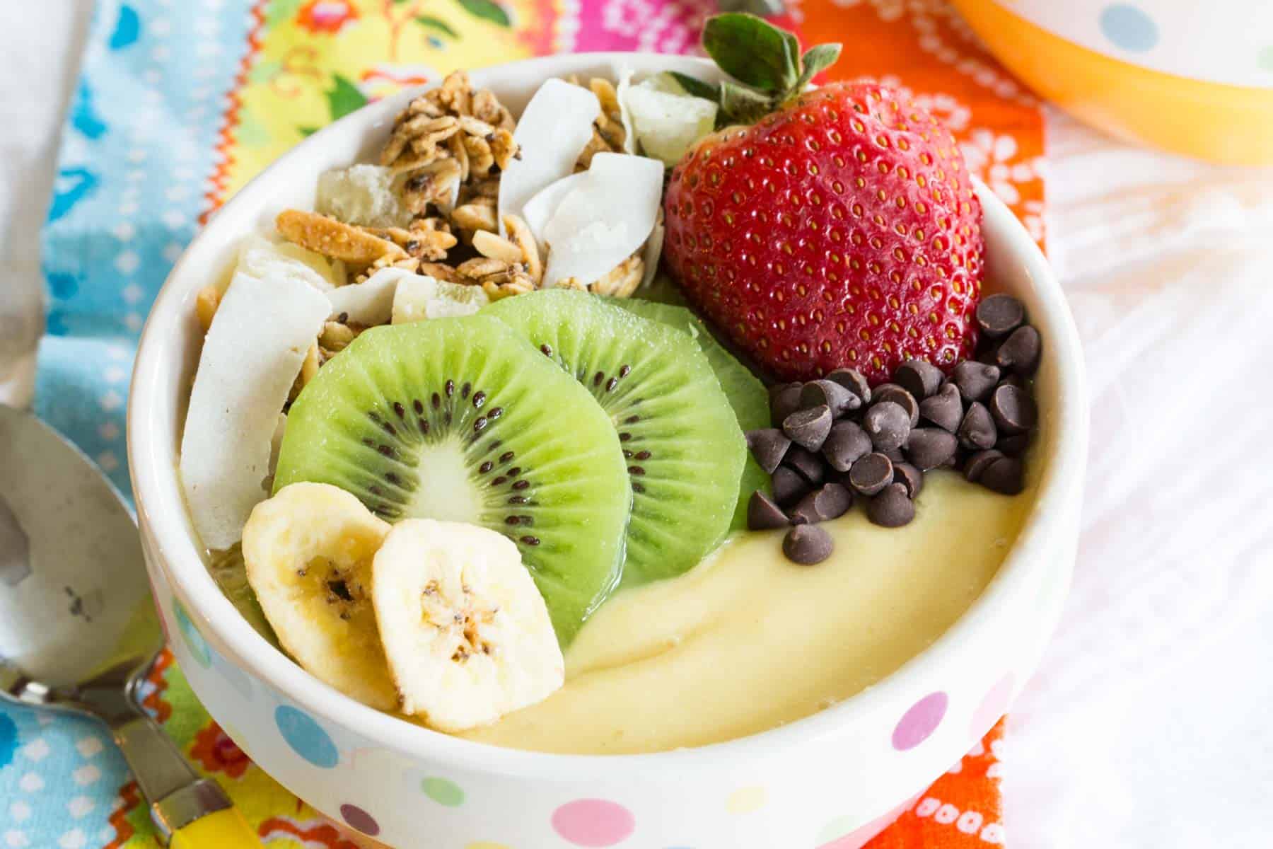 pink and white polka dot bowl filled with a thick pale yellow smoothie and fruit and granola on top