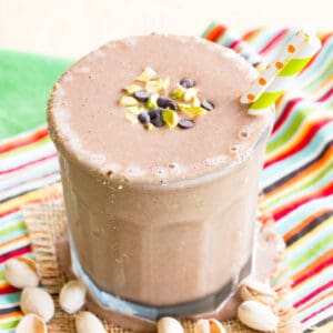 A chocolate smoothie in a glass topped with chopped pistachios and mini chocolate chips on top of cloth napkins.
