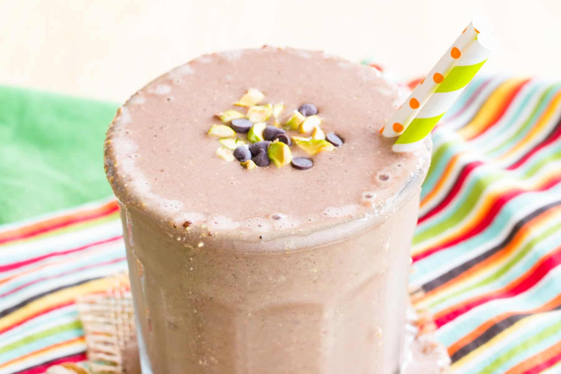 Looking at the top of a chocolate smoothie in a glass with two straws and some chopped pistachios and mini chocolate chips on top.