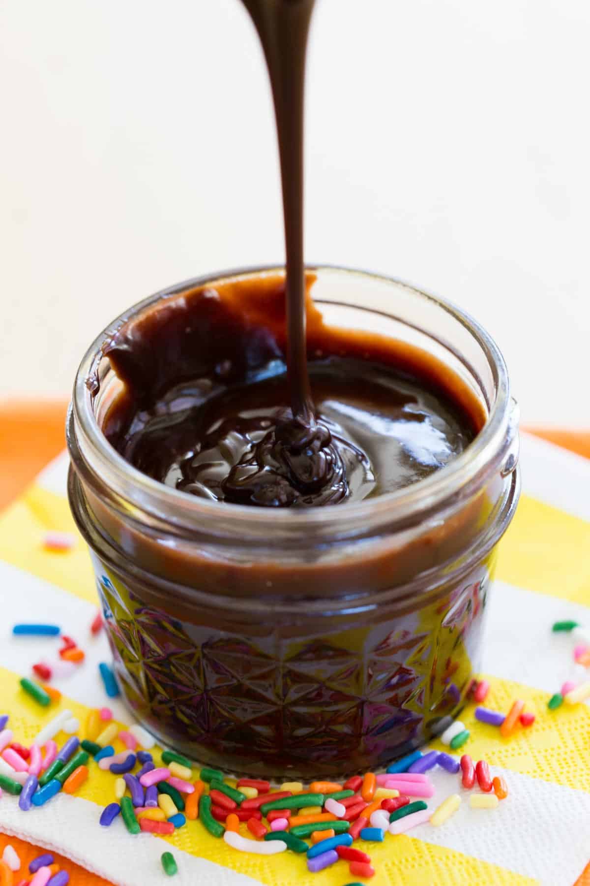 hot fudge sauce dripping into a small jar on a yellow and white striped napkin surrounded by rainbow sprinkles