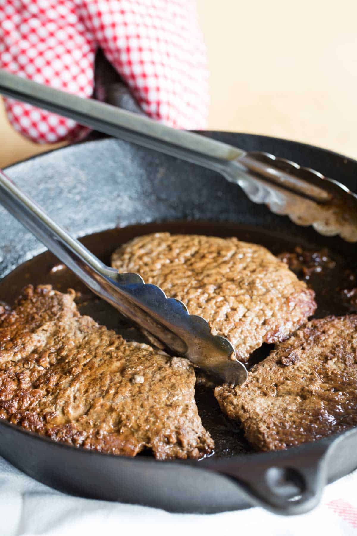 Three cubed steaks in a cast iron skillet with a set of metal tongs.