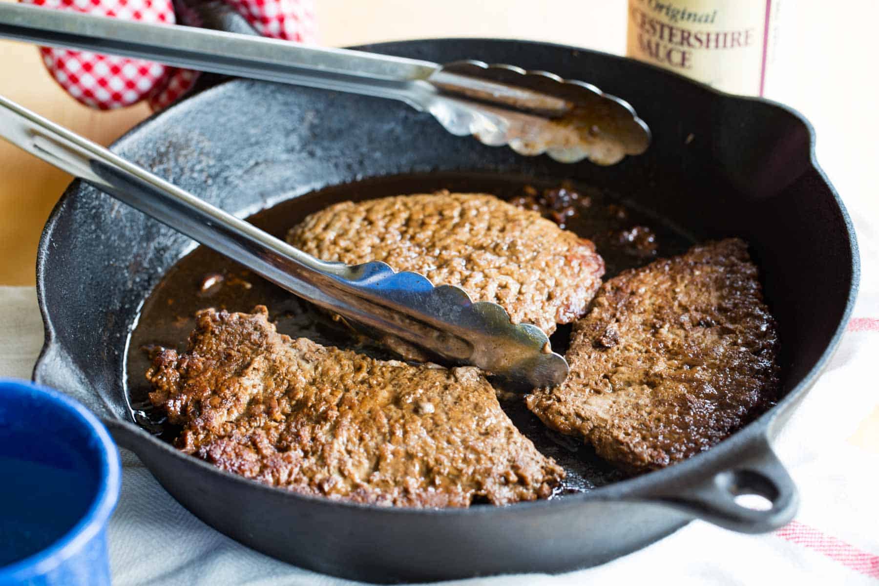 Three cube steaks in a cast iron skillet with a set of tongs.