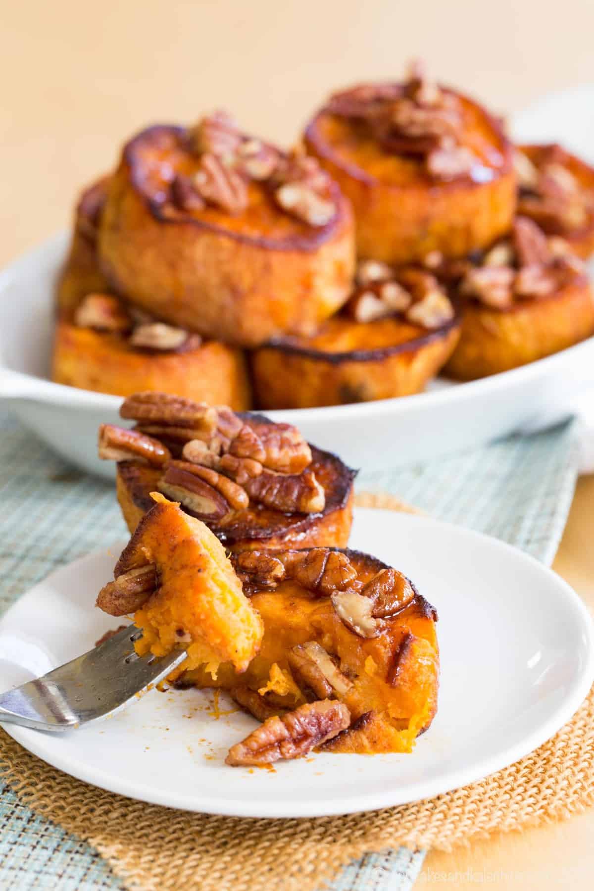 A forkful of sweet potato in the foreground with a dish of melting potatoes topped with pecans in the background