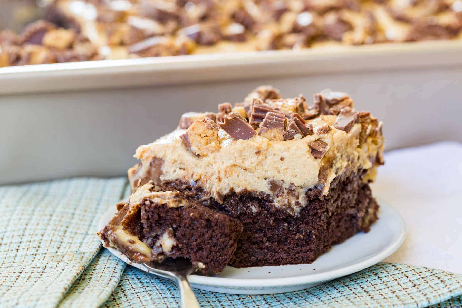 Apiece of Reeses Peanut Butter Cup Poke Cake with a fork on a small white plate on top of a blue plaid cloth napkin.
