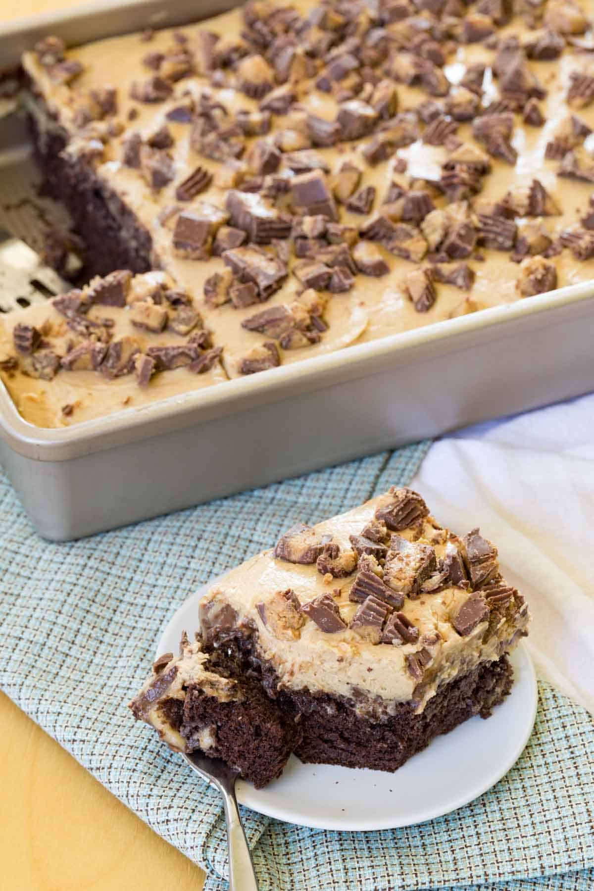 A piece of Reese's poke cake topped with peanut butter cups on a plate next to the cake pan with the rest of the cake.