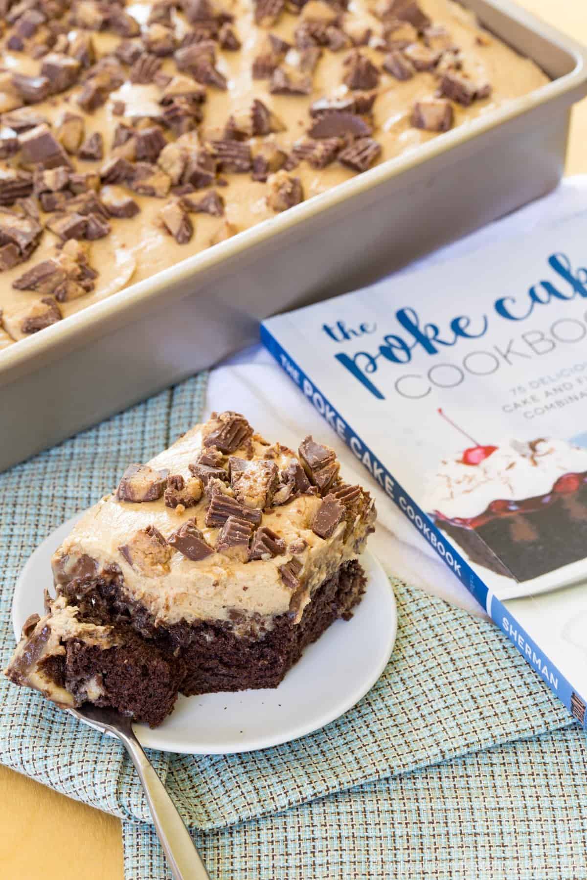 A piece of reese's cake on a plate next to the rest of the cake in a pan and a copy of The Poke Cake Cookbook.