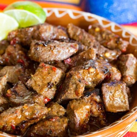 A terra cotta dish of steak bites with lime wedges on a striped cloth napkin.