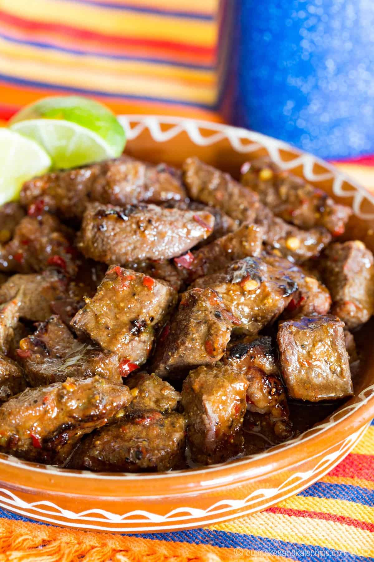 A terra cotta dish of steak bites with lime wedges on a striped cloth napkin.