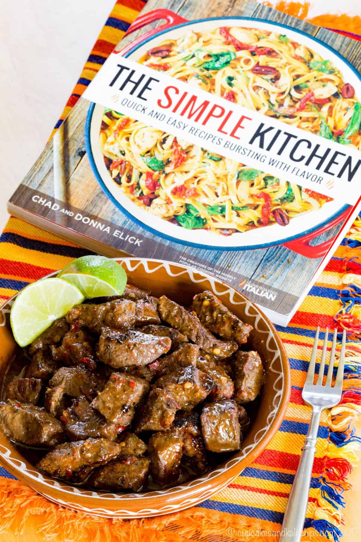 A dish of Chili Lime Steak Bites and The Simple Kitchen Cookbook on top of a striped napkin.