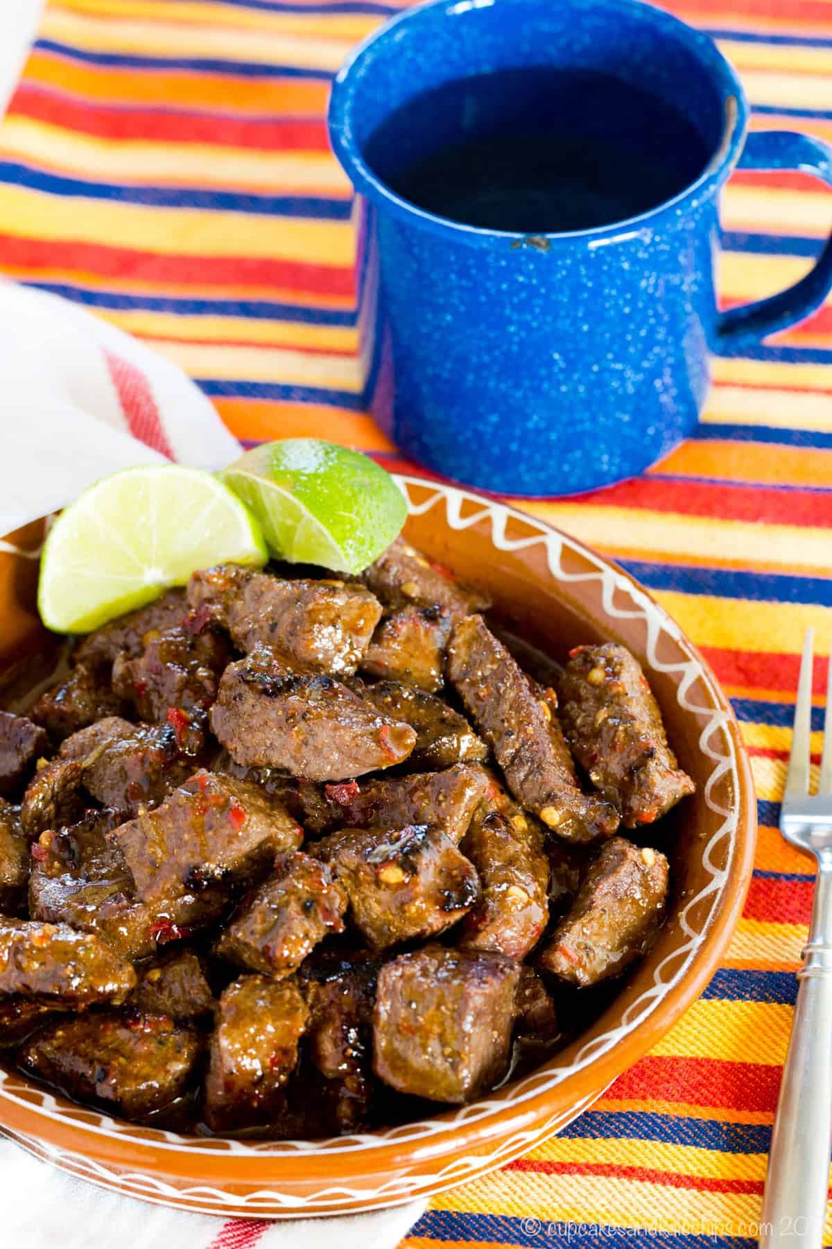 Steak Bites coated in a glaze in a dish with lime wedges next to a blue speckled cup of water.