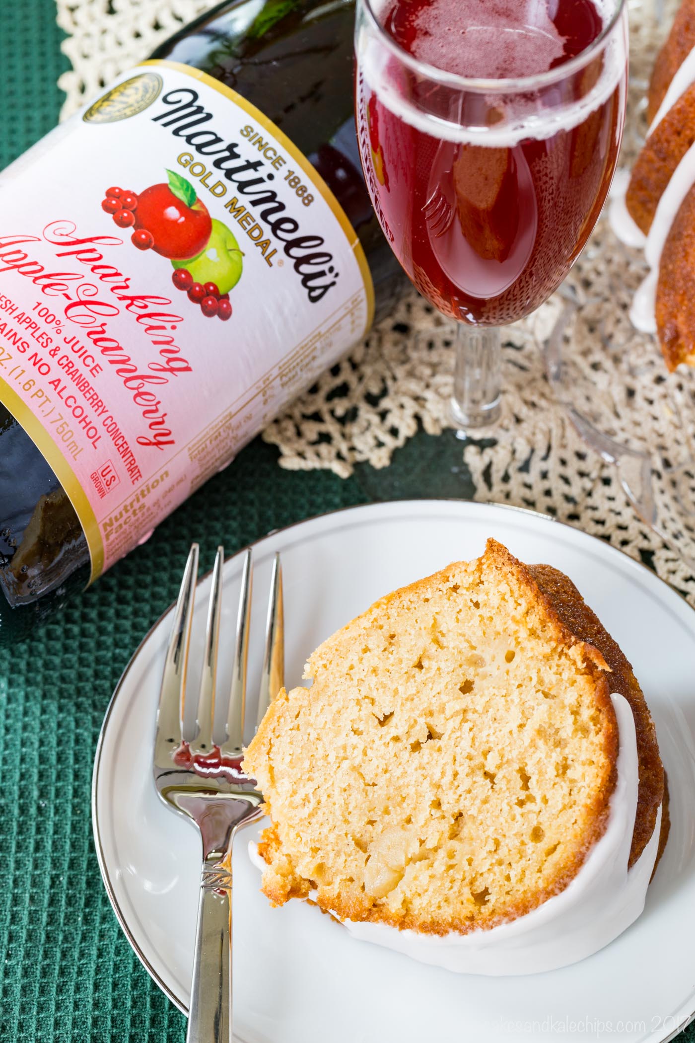 A slice of gluten-free apple cranberry donut cake on a plate next to a fork and a bottle of Martinelli's sparkling cider.