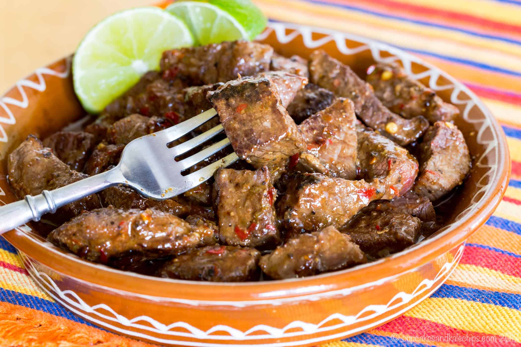 A fork laying in a bowl of chili lime steak bites with two lime wedges.