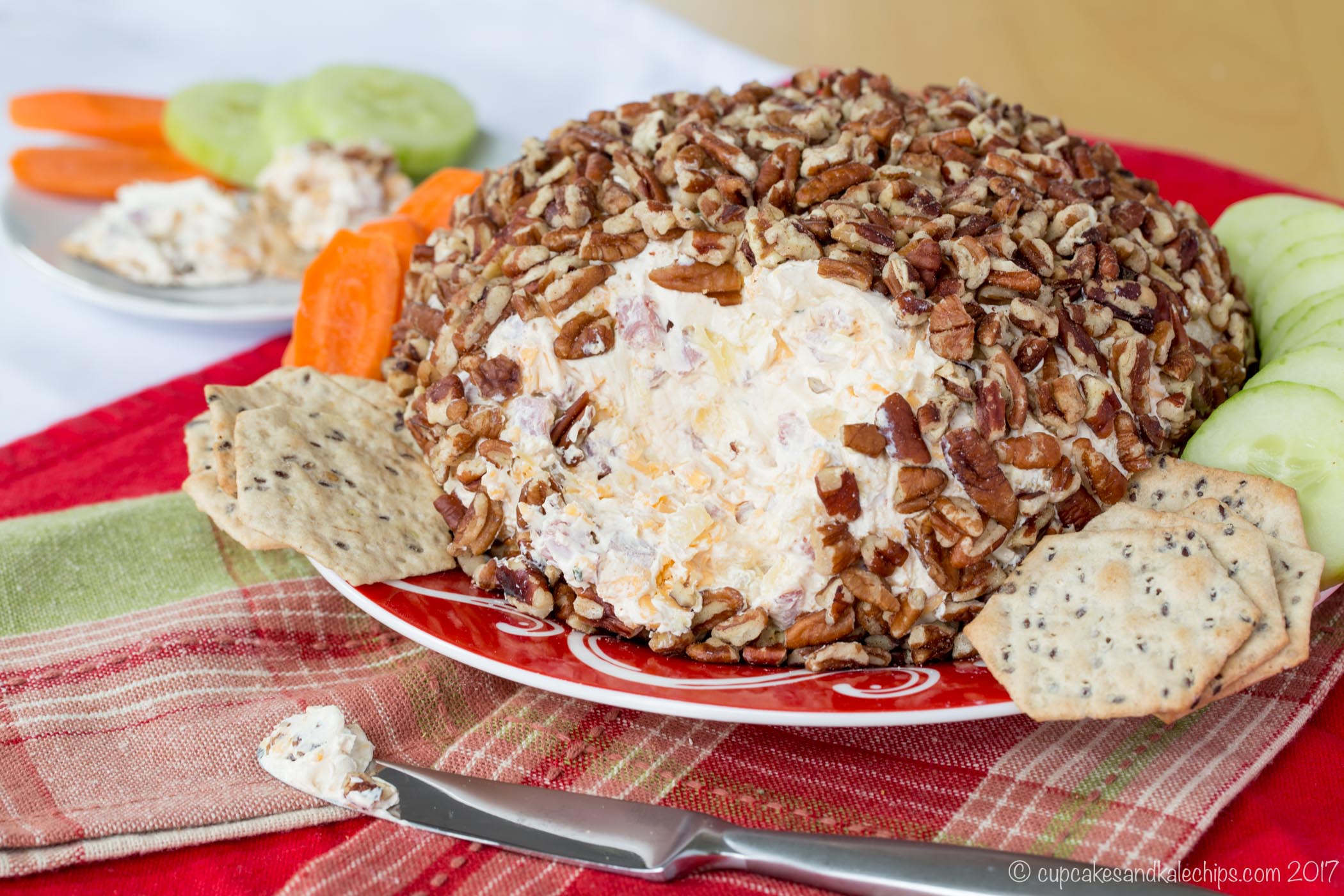 A pineapple and ham cheeseball on a red and white plate with crackers and veggies set on top of red and plaid cloth napkins on a table.