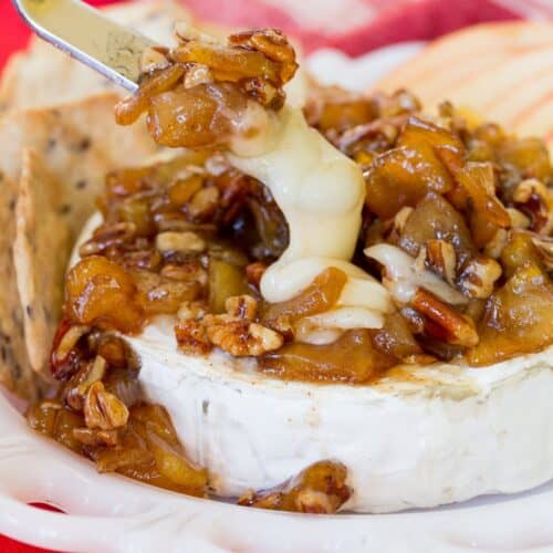 A spreading knife scooping up melty Baked Brie topped with appled and pecans.