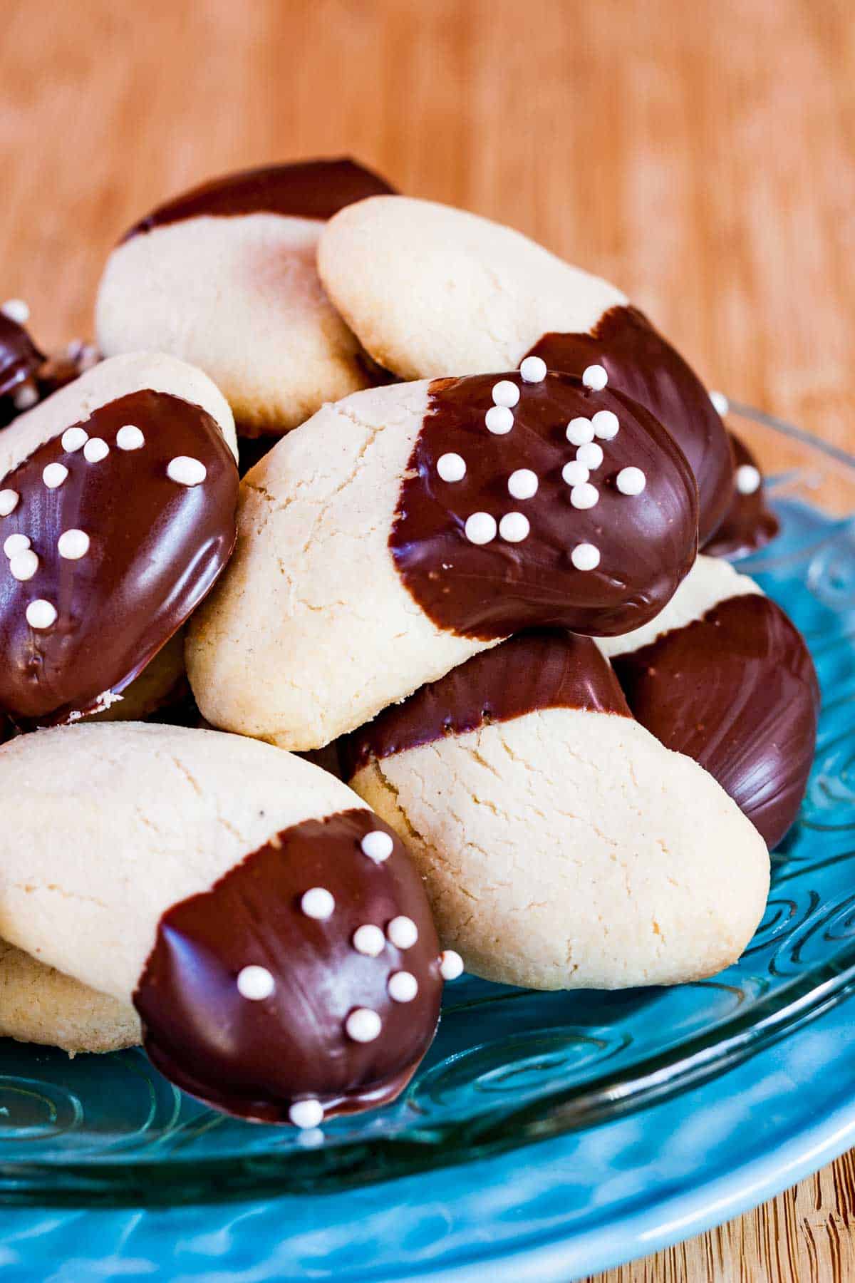 A pile of chocolate-dipped gluten free almond crescent cookies on a glass plate set on top of a light blue dish.