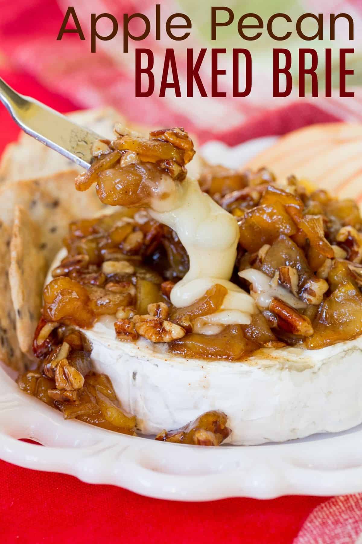 A wheel of baked brie cheese in the foreground topped with a caramelized apple pecan topping, with a bottle of sparkling cider in the background