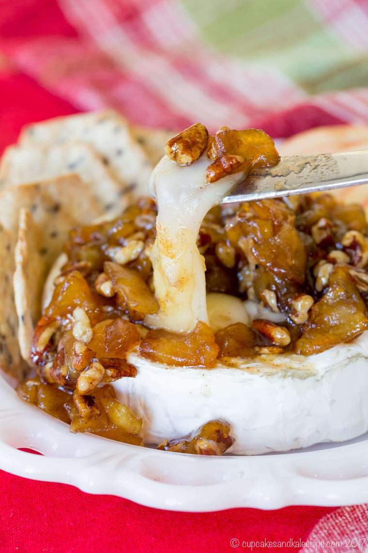 A cheese knife cuts into a melted wheel of Caramelized Apple Pecan Baked Brie