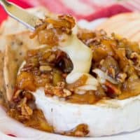 A cheese knife cuts into a melted wheel of Caramelized Apple Pecan Baked Brie