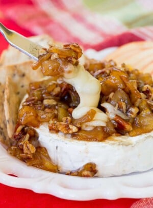 A cheese knife cuts into a melted wheel of Caramelized Apple Pecan Baked Brie