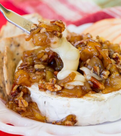 A cheese knife cuts into a melted wheel of Caramelized Apple Pecan Baked Brie