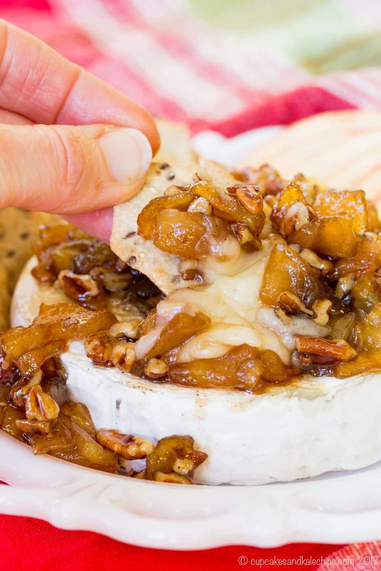 A hand dipping a cracker into the melted cheese of this baked brie with caramelized apples and pecans
