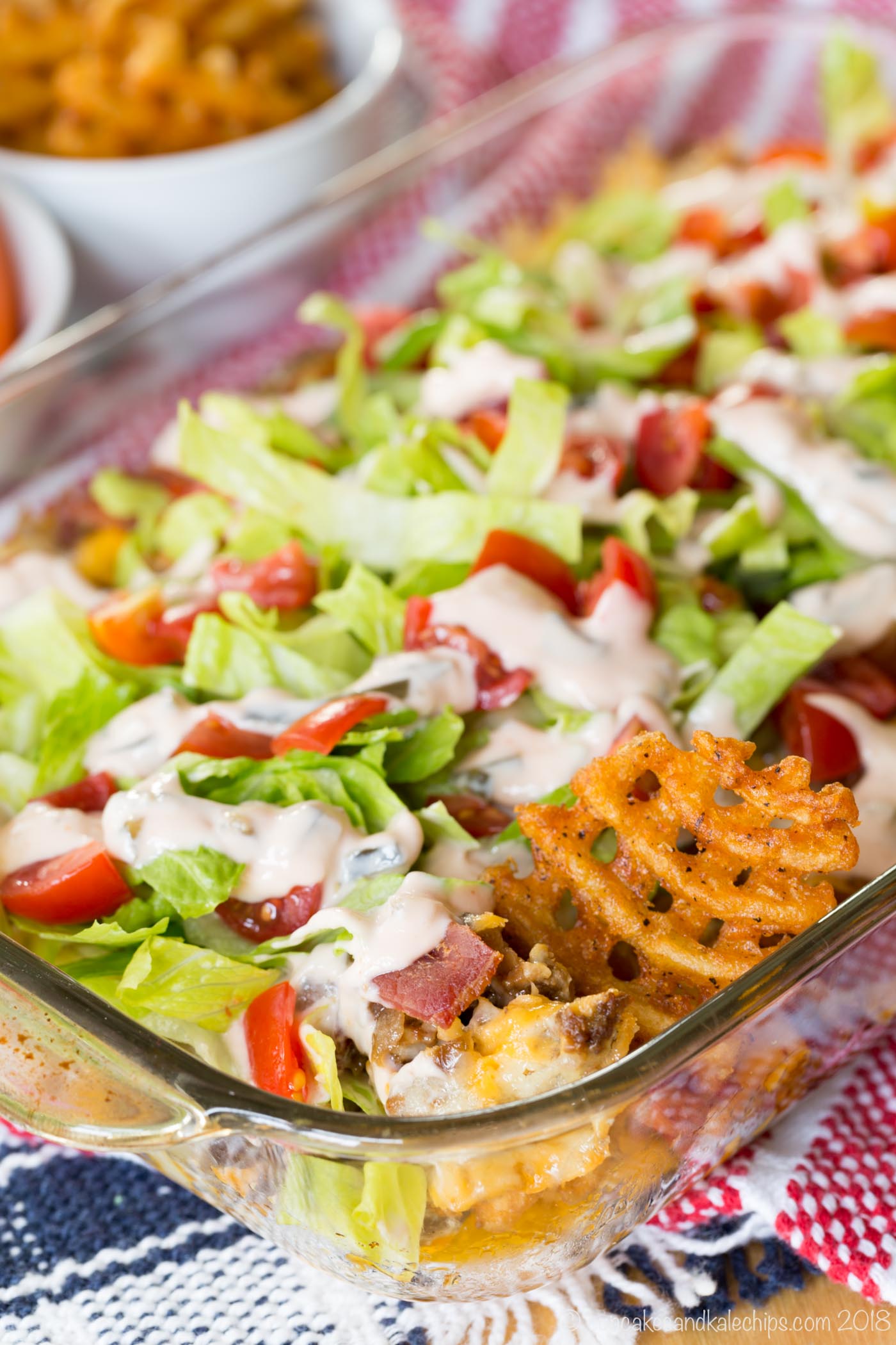 A waffle fry stuck in a pan of bacon cheeseburger dip topped with lettuce, tomato, and a drizzle of sauce.