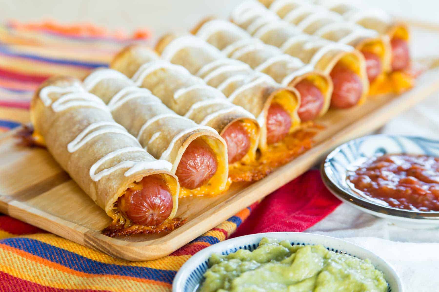 A row of Mexican Hot Dog Taquitos on a wooden serving plate with small dishes of salsa and guacamole.