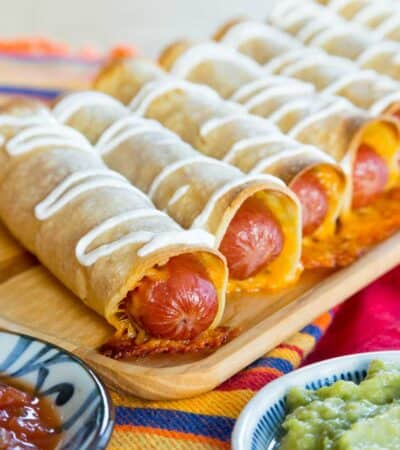 Crispy Hot Dog Taquitos drizzled with sour cream and lined up on a wooden platter.