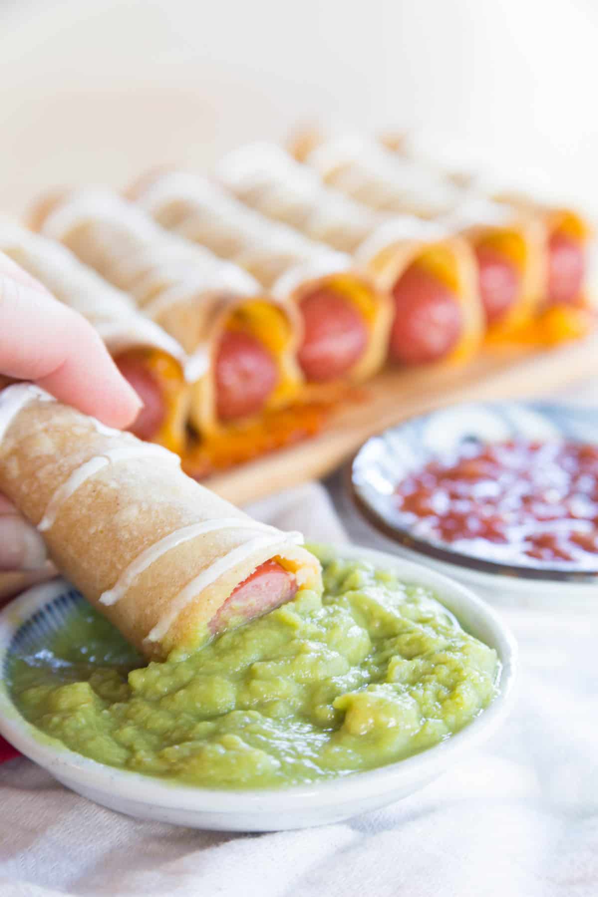 A hot sog wrapped in a crispy tortilla being dipped in Guacamole.