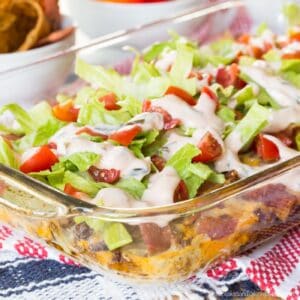 Looking at the side of a glass baking dish with a layered Cheeseburger Dip with pieces of bacon, shredded lettuce, chopped tomatoes, and a creamy sauce.