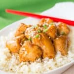 Chunks of orange chicken over cauliflower rice in a white oval dish with red chopsticks resting on it.