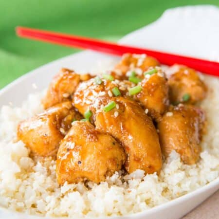 Chunks of orange chicken over cauliflower rice in a white oval dish with red chopsticks resting on it.