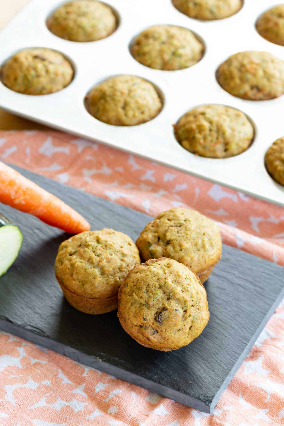 three gluten free mini muffins on a platter in front of a pan of more carrot zucchini muffins