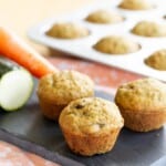 a pan of gluten free mini carrot zucchini muffins with three on a slate tray in front of it