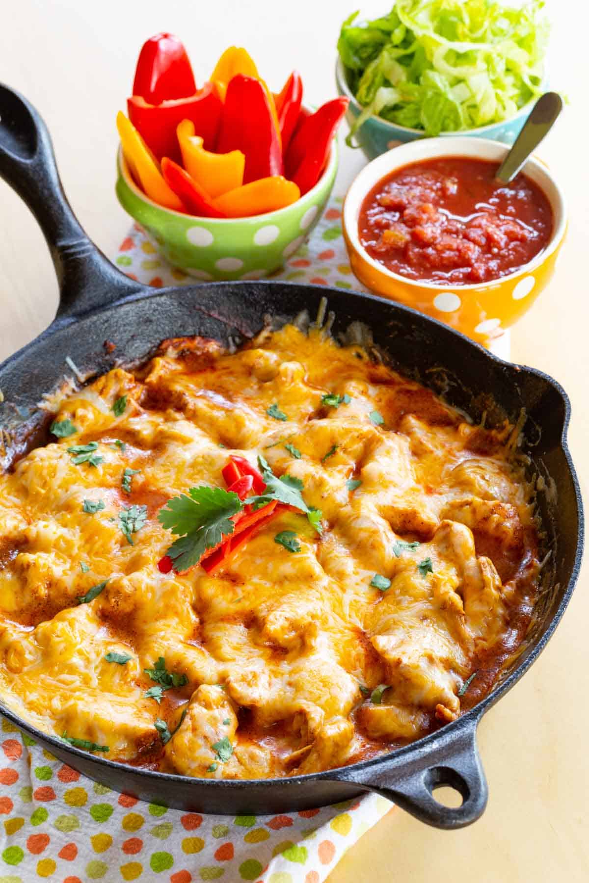 cast iron pan with the chicken enchilada skillet recipe cooked in it and topped with melted cheese next to small bowls of lettuce, salasa, and mini bell peppers