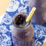Two blueberry smoothies in glass bottles on top of a blue cloth napkins with white flowers.