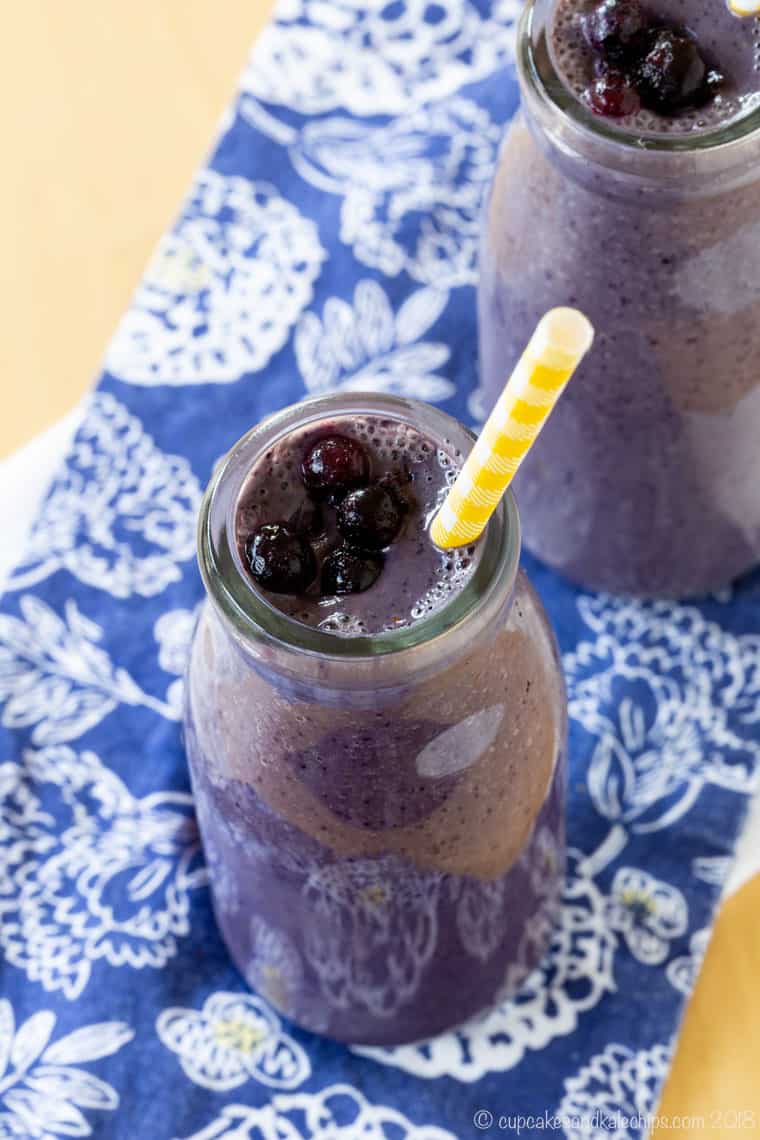 Two blueberry smoothies in glass bottles on top of a blue cloth napkins with white flowers.