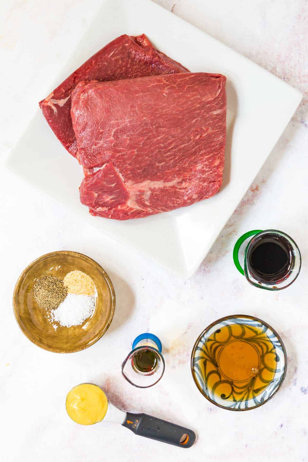 raw steaks on a plate with containers of balsamic vinegar, olive oil, honey, dijon mustard, and spices on a marble tabletop