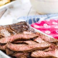 sliced grilled flat iron steak on a white serving plate with bowls of side dishes behind it