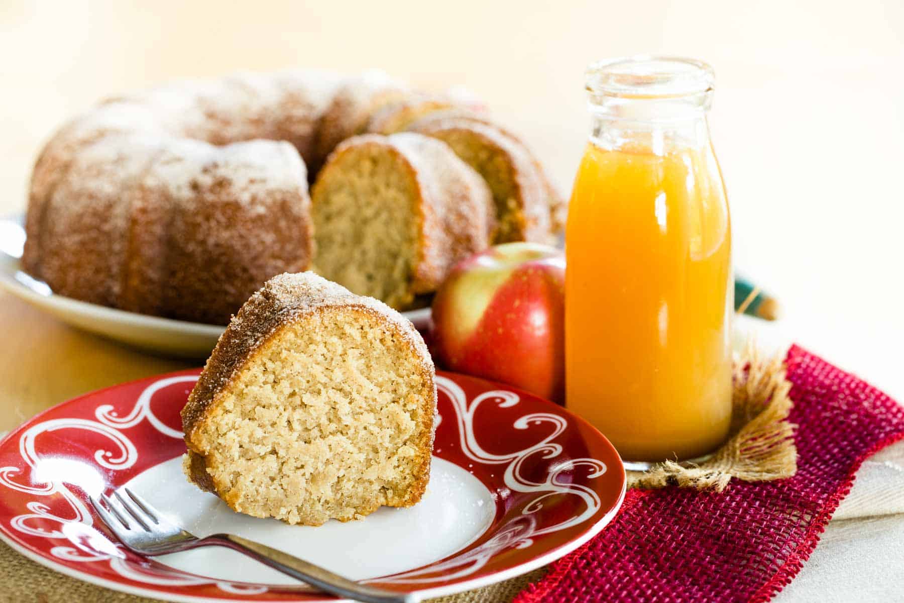 Slice of gluten free apple cider donut cake on a plate with a red and white design and the rest of the bundt cake on a serving plate behind it plus a bottle of apple cider