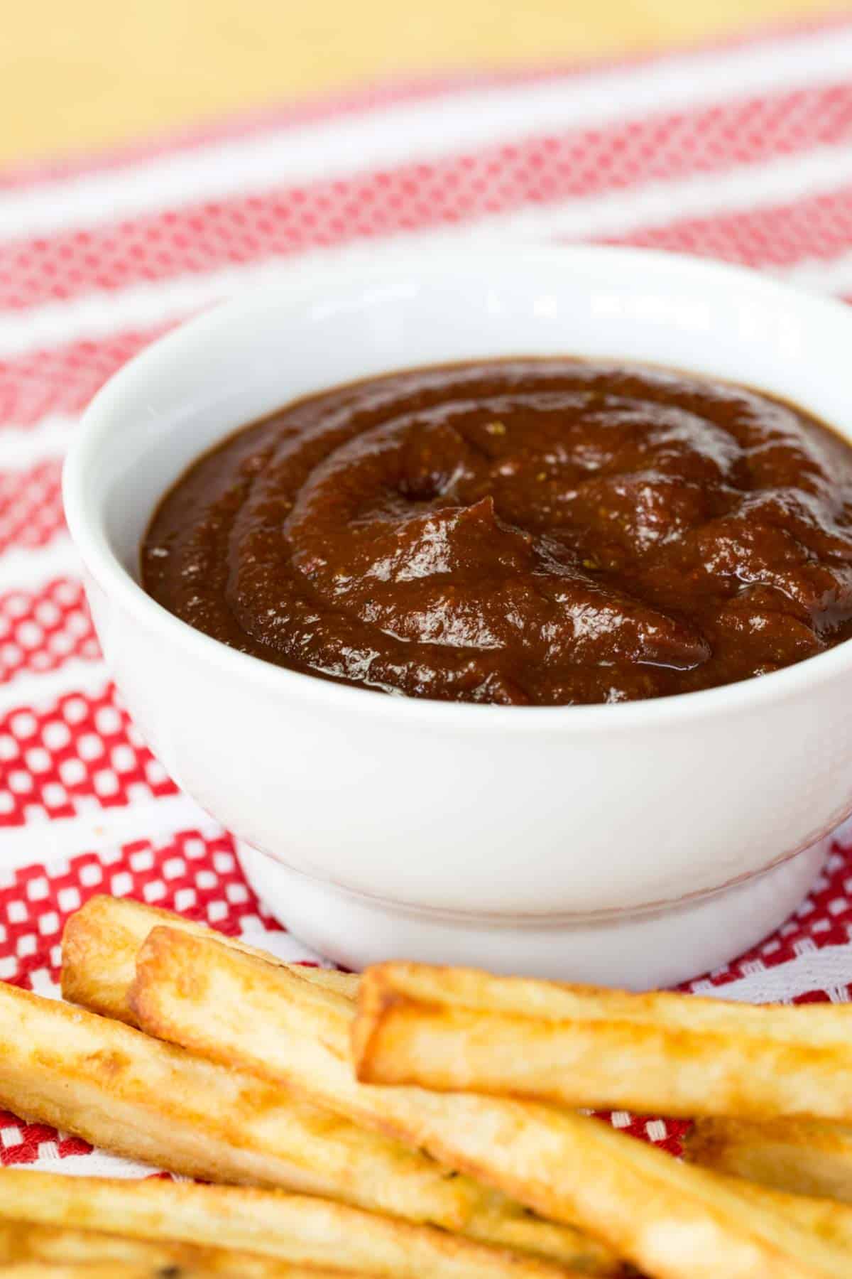 A white bowl of balsamic ketchup on a red and white napkin with some fries in front of it.