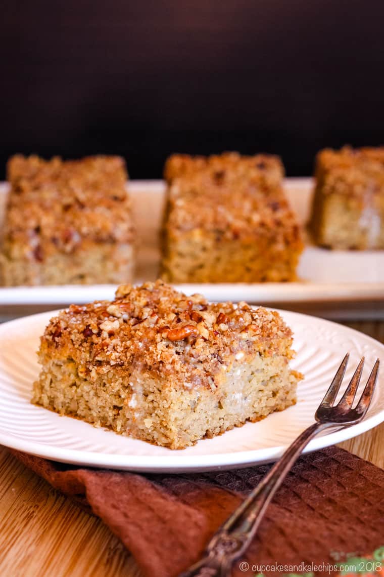 A square piece of pumpkin coffee cake on a plate with a small dessert fork and more pieces on a platter behind it.