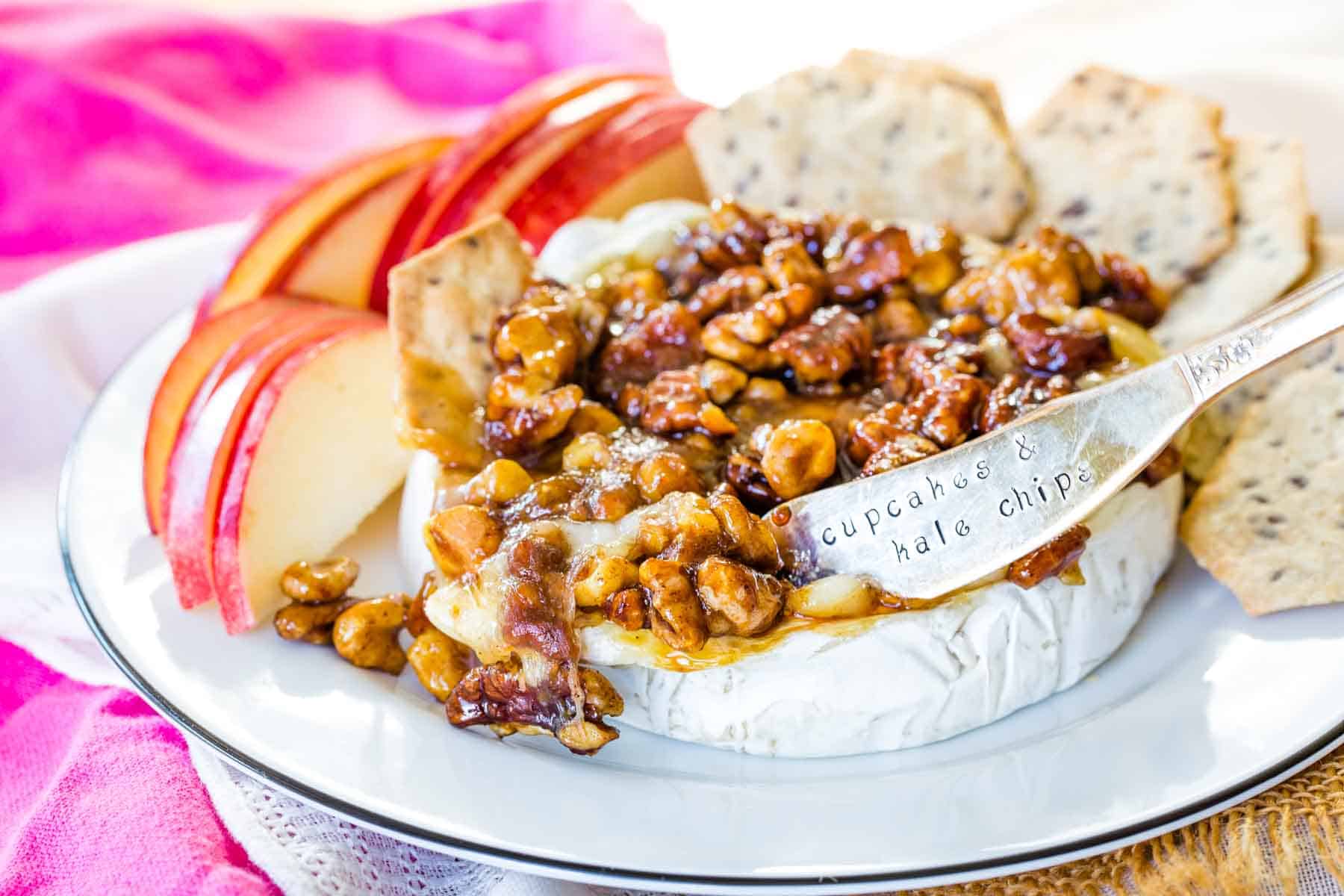 Honey Nut Baked Brie Cheese on a small plate with apple sliced and crackers with a silver spreader knife in it.