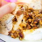 A cracker being dipped into baked brie topped with honey-glazed nuts.