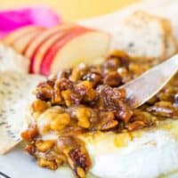 A small spreader starting to scoop up some melty baked brie topped with honey-glazed walnuts and pecans.