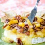 A spreader knife in a wheel of melty Brie on a plate with text overlay that says "Pineapple Pecan Baked Brie".