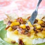 A small spreader knife in a melty wheel of Brie topped with pineapples and pecans.