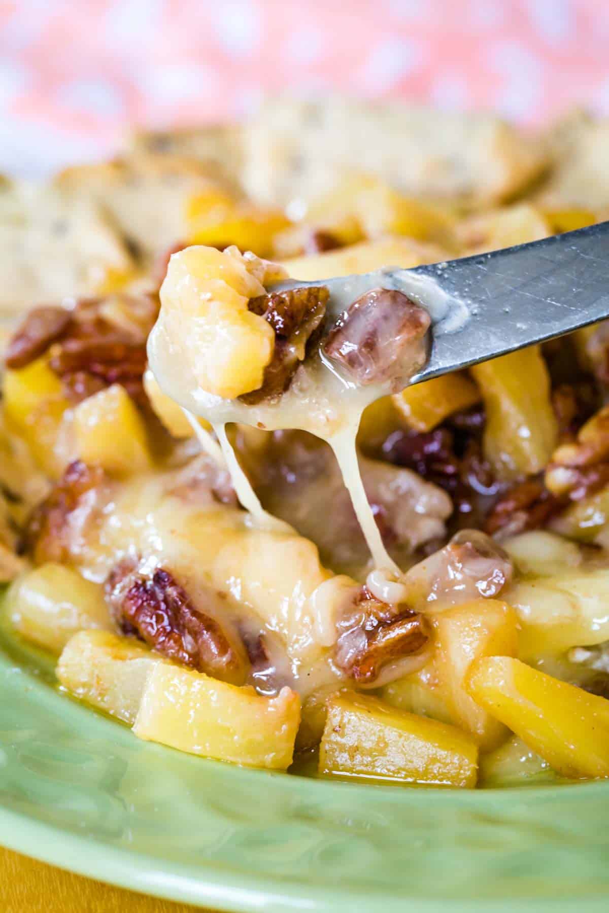 A small spreader knife with melty baked brie dripping off of it over the top of the round of cheese with pineapple and pecans.