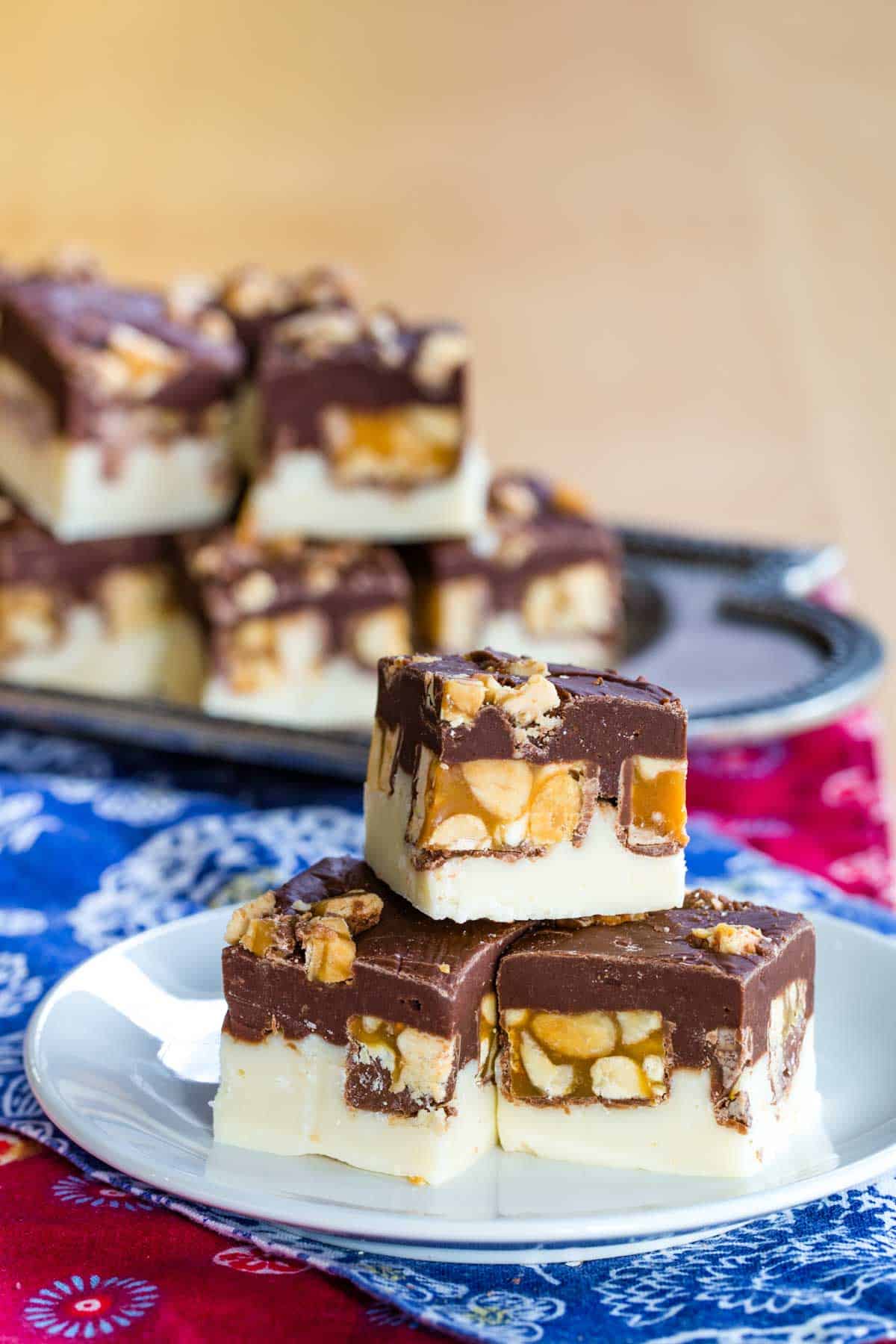 Small white plate with three squares of Double Chocolate Snickers Fudge on top of floral cloth napkins with a silver platter of more fudge in the background.